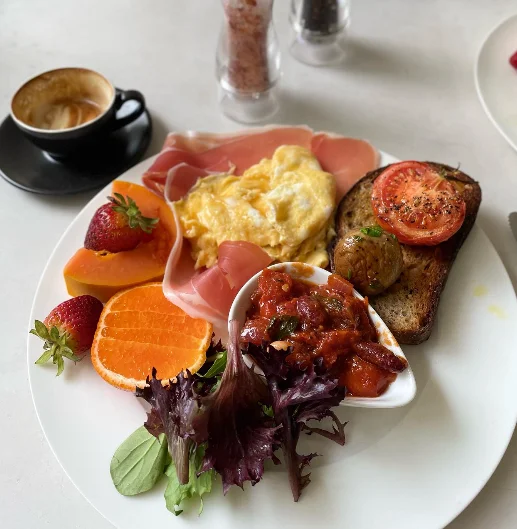 Breakfast plate of scrambled eggs, prosciutto, sourdough with roast tomato and mushroom, beans, salad and fresh fruit, with a coffee