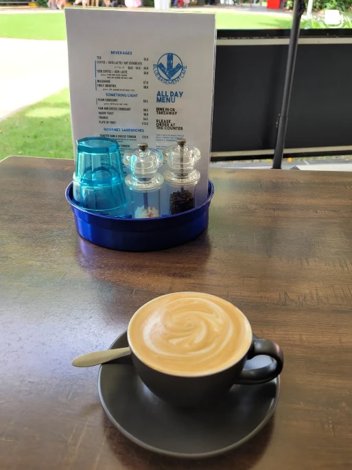 Flat white with latte art on a timber table, with the Blue Basement Cafe all day menu behind