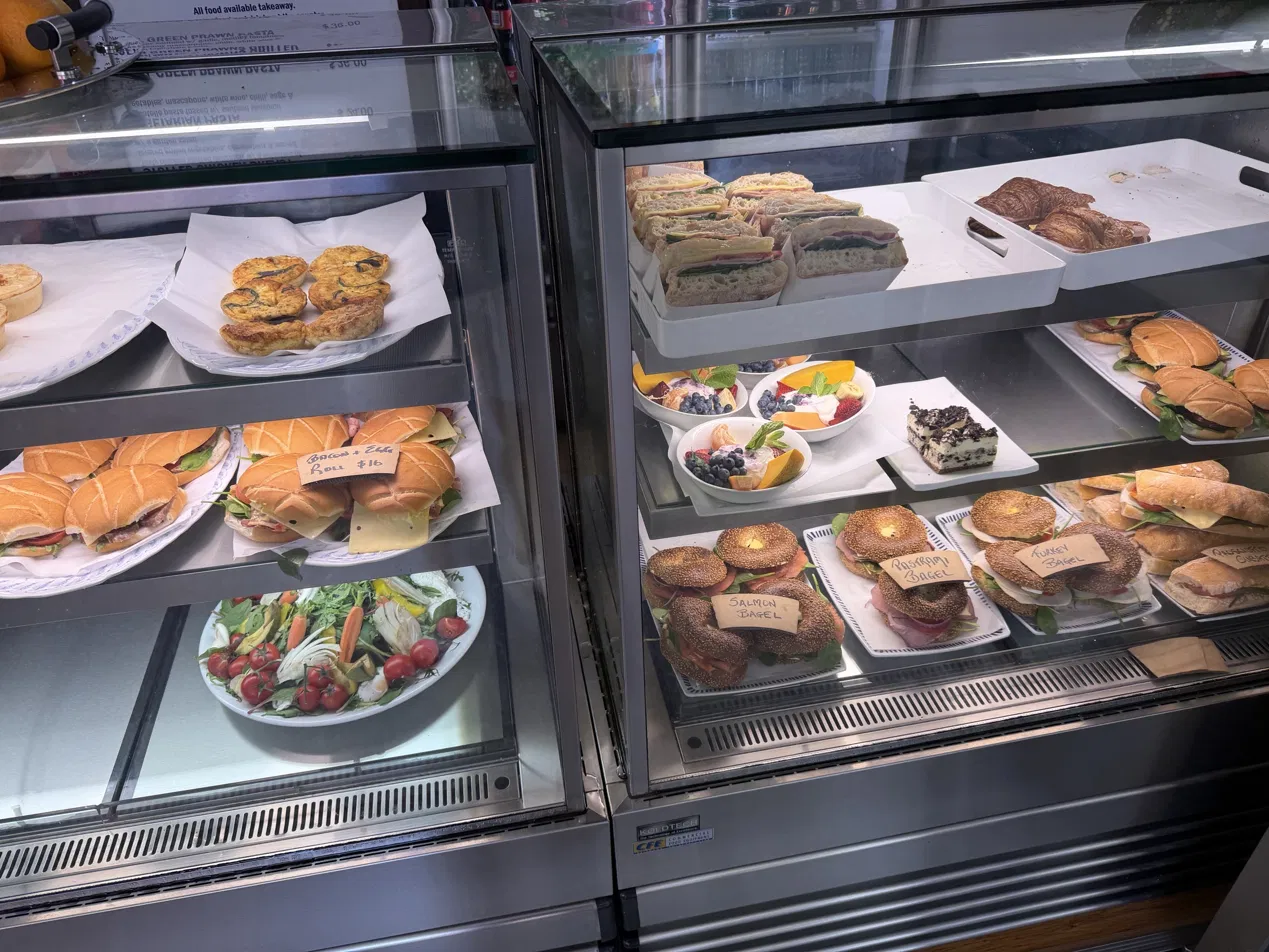 Food cabinet stocked with salmon, pastrami and turkey bagels, bacon and egg rolls, sandwiches, quiches, fruit bowls and salad